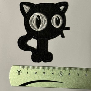 Op de afbeelding: Zwarte geborduurde strijkpatch van een cartoonkat met grote witte ogen. De kat staat op twee poten en heeft een staart.