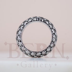 Peut inclure: Une bague argentée avec un motif texturé en forme d'écailles. La bague a une large bande et un design détaillé qui enveloppe toute la circonférence. La bague est présentée sur un fond doux et neutre.