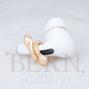 Puede incluir: Un anillo abstracto de color dorado en forma de serpiente. El anillo está encima de un auricular inalámbrico blanco.