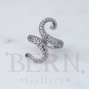 May include: A silver ring shaped like an octopus tentacle. The ring features detailed suckers and a curled end. The design wraps around the finger. The background is white.