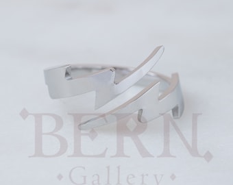 May include: A silver-colored ring with a lightning bolt design. The ring features two stylized lightning bolts that wrap around the finger. The ring is made of a shiny, reflective metal and is set against a white background.