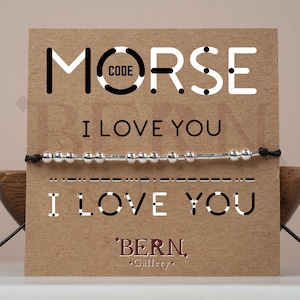 May include: A brown card with the words "MORSE CODE" and "I LOVE YOU" in black text. A silver beaded bracelet with a black cord is displayed on the card. The bracelet is a Morse code bracelet that spells out "I LOVE YOU". The card also has the text "BERN Gallery" in black text.