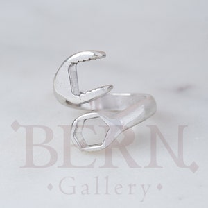 May include: Silver adjustable ring shaped like a wrench. The ring is open on one side and has a hexagonal shape on the other side.