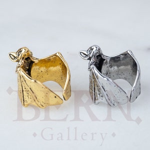May include: Two bat-shaped rings, one gold and one silver, with detailed wings and a bat head on the top. The rings are open on one side and can be adjusted to fit different finger sizes.