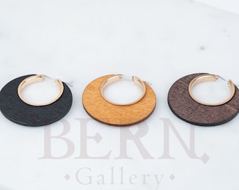 Puede incluir: Tres pendientes de aro de madera en tonos marrones y beige. Cada pendiente presenta un aro interior dorado. Los pendientes están dispuestos sobre una superficie blanca, mostrando sus colores y texturas contrastantes.
