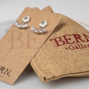 May include: A pair of silver earrings with white flower accents and clear rhinestones. The earrings are displayed on a brown card with the text "BERN Gallery".