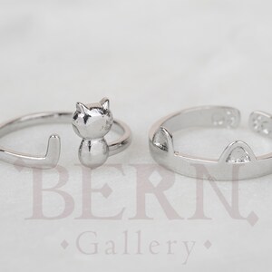 Op de afbeelding: Twee zilveren ringen met kattenontwerpen. Een ring heeft een katten silhouet met een staart die om de vinger wikkelt. De andere ring heeft kattenoren aan de bovenkant.