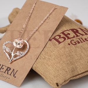May include: Rose gold necklace with a heart-shaped pendant featuring a rose and sparkling crystals. The necklace is displayed on a brown card with the "BERN Gallery" logo, next to a burlap bag.