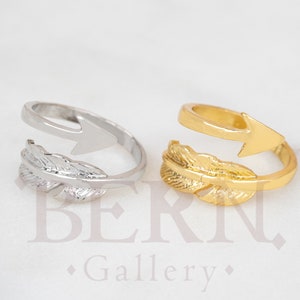 Könnte beinhalten: Zwei verstellbare Ringe mit einem Feder- und Pfeil-Design. Ein Ring ist silber und der andere ist gold.
