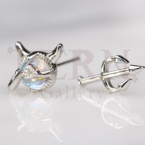 May include: A pair of silver earrings, one featuring a moonstone set in a silver devil's head design, and the other featuring a silver trident.