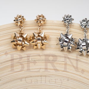 May include: A pair of gold and silver bow-shaped earrings. The earrings are dangling and have a three-dimensional bow shape.