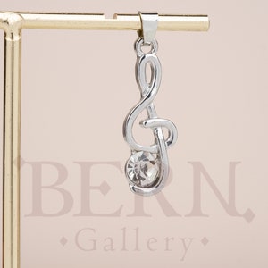 Puede incluir: Encanto de clave de sol de plata con una piedra de cristal transparente. El encanto cuelga de un soporte dorado.