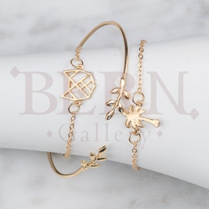 May include: Gold-toned bracelet set with three charms: a geometric design, a palm tree, and a leafy branch. The bracelet is made of a thin chain and a thicker bangle.