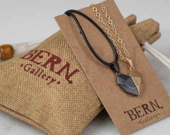 May include: Two necklaces with heart-shaped pendants. One necklace has a black leather cord and a black and gold heart pendant. The other necklace has a gold chain and a gold heart pendant. The necklaces are displayed on a brown card with the text "BERN Gallery."