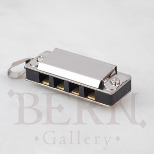 May include: A small silver harmonica with black and gold detailing. The miniature musical instrument has a rectangular shape and a loop for attaching to a keychain or necklace. The harmonica is a musical instrument.