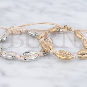 Könnte beinhalten: Zwei Armbänder mit einem beigefarbenen Band und silbernen und goldenen Muschel-Anhängern.