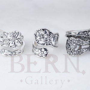 Puede incluir: Tres anillos de plata con forma de cuchara y diseños florales se muestran sobre un fondo blanco. Cada anillo está hecho de una cuchara, con la parte cóncava de la cuchara formando la cara del anillo. Los anillos muestran detalles y texturas.