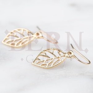 May include: A pair of gold-coloured leaf-shaped earrings. Each earring has a delicate, openwork design with the veins of the leaf visible. The earrings hang from simple, curved ear wires, showcasing a minimalist and elegant style.