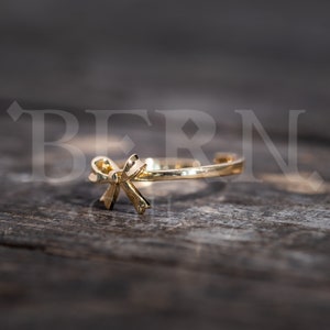 May include: A gold-coloured ring with a bow design. The ring is lying on a wooden surface.