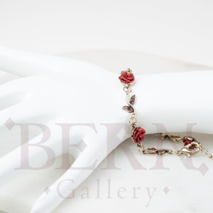 May include: A gold-toned bracelet featuring red rose accents and small red gems. The bracelet is designed with a delicate chain and is displayed on a white mannequin hand. The roses are a vibrant red color.