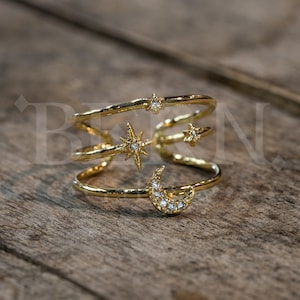 May include: Gold-toned ring featuring celestial designs. The ring has a layered design with a crescent moon, stars, and a starburst. The moon and stars are embellished with small, clear gemstones. The ring is a unique piece of jewelry.