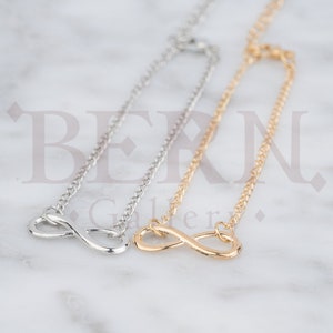 May include: Two infinity symbol necklaces, one silver and one gold, on a white marble background.