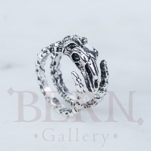 May include: A silver ring featuring a detailed skull design. The ring is made of two bands that are intertwined.