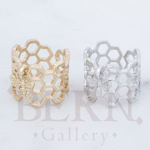 Könnte beinhalten: Zwei verstellbare Ringe mit einem Wabenmuster und einem Bienen-Detail. Ein Ring ist golden und der andere ist silber.
