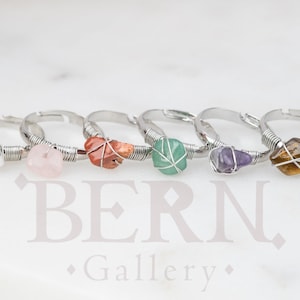 May include: Six silver rings with different coloured stones wrapped in wire. The stones are white, pink, red, green, purple, and brown.