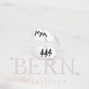 Può includere: Anello d'argento con un design a forma di montagna e albero. L'anello è aperto e regolabile.