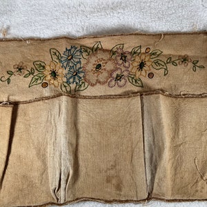 May include: A tan fabric organizer with three pockets. The top section features embroidered flowers in blue, yellow, and purple with green leaves. The edges are frayed, giving it a rustic, vintage appearance. Ideal for wall storage.