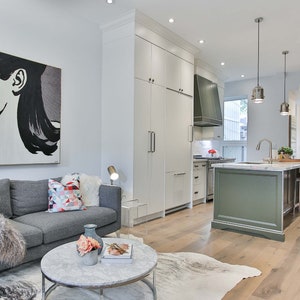 May include: A modern living room with a gray sofa, a white fur throw, and a coffee table with a marble top. The room has a large pop art print on the wall and a view of a kitchen with green cabinets.