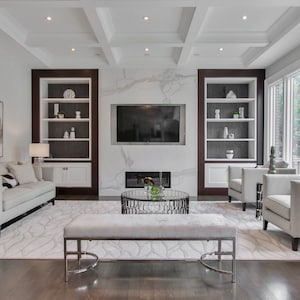 May include: A modern living room with a neutral color palette. Features include a white sofa, two armchairs, a bench, and a coffee table. Built-in bookshelves flank a fireplace and a large TV. The room has hardwood floors and large windows.