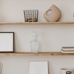 May include: A wooden shelf unit with three shelves. The top shelf has a brown candle, a black wire basket with round objects, and a beige ceramic vase. The middle shelf has a black framed picture and a clear glass decanter. The bottom shelf has a set of black incense sticks, a white framed picture, and a white mug with a black design.