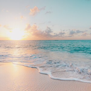 May include: A sandy beach with turquoise water lapping at the shore. The sun is setting over the horizon, casting a warm glow on the scene.