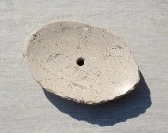 May include: A light beige, oval-shaped stone soap dish with a central drainage hole. The dish has a rough, textured surface and a natural, organic aesthetic. The soap dish is designed to hold a bar of soap.