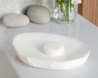 May include: A white, oval-shaped soap dish with a textured surface. A round, white bar of soap rests in the center. The dish is on a white surface, with two smooth, gray stones and a vase of flowers in the background.