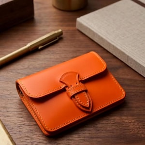 Pode incluir: Carteira de couro laranja com fecho de aba e fivela decorativa. A carteira retangular tem costuras visíveis. Uma caneta dourada e um caderno são visíveis no fundo.