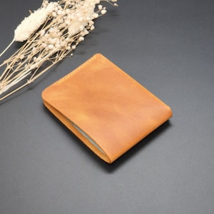 May include: A tan leather wallet with a minimalist design. The wallet is folded and lying on a dark gray surface.