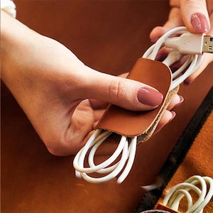 May include: A brown leather cable organizer holding white USB cables. The organizer is a small, rectangular piece of leather with a slot to hold the cables. The cables are neatly coiled and ready for use. The background is a warm brown color.