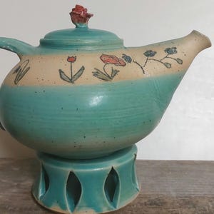 Könnte beinhalten: Eine teelfarbene Keramik-Teekanne mit Blumenmuster und einem passenden Ständer. Die Teekanne hat einen runden Körper und einen Ausguss an der Seite. Der Ständer hat ein offenes Design mit Ausschnitten.