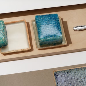 Könnte beinhalten: Eine Sammlung von Keramik-Seifenschalen und ein Buttermesser. Die Seifenschalen sind quadratisch und rechteckig, mit türkisfarbenen und weißen floralen und geometrischen Mustern. Das Messer hat eine silberne Klinge und einen dekorativen Griff.