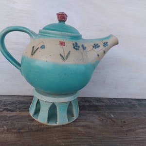 Könnte beinhalten: Eine türkisfarbene Teekanne mit weißem Sockel und Blumendesigns. Die Teekanne hat einen roten Knopf auf dem Deckel und steht auf einem passenden türkisfarbenen Ständer mit Ausschnitten.