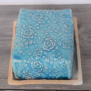 Könnte beinhalten: Eine blaue Keramik-Butterdose mit einem weißen Blumenmuster. Die Dose hat einen erhöhten Rand und eine glatte, glasierte Oberfläche.