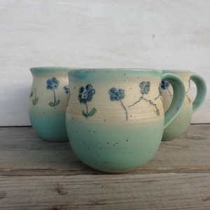 Könnte beinhalten: Drei handgefertigte Keramikbecher mit helltürkisfarbenem Boden und cremefarbenem Oberteil. Jeder Becher hat ein handgemaltes blaues Blumenmuster. Die Becher werden auf einer verwitterten Holzoberfläche präsentiert, was ihre handwerkliche Qualität hervorhebt.