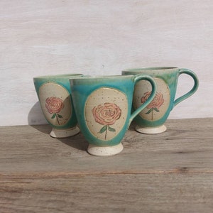 Könnte beinhalten: Drei Keramikbecher mit türkisfarbener Glasur und cremefarbenem Sockel. Jeder Becher zeigt ein rosafarbenes Rosendesign in einem cremefarbenen Oval. Die Becher haben einen Henkel und stehen auf einer Holzoberfläche.
