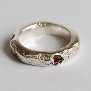 Może przedstawiać: Srebrny pierścionek z fakturowaną powierzchnią i małym, ciemnoczerwonym kamieniem szlachetnym.