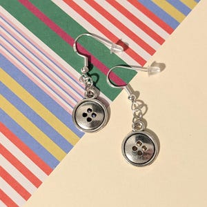 May include: A pair of silver button earrings. Each earring has a small, round, silver button charm with four holes. The earrings hang from silver fish hook ear wires. The background is a colourful striped pattern.