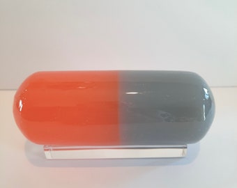 May include: A large, orange and gray glass pill shaped sculpture on a clear acrylic stand.