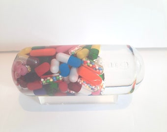 May include: A clear glass bottle filled with colorful pills and capsules. The bottle is on a white stand.